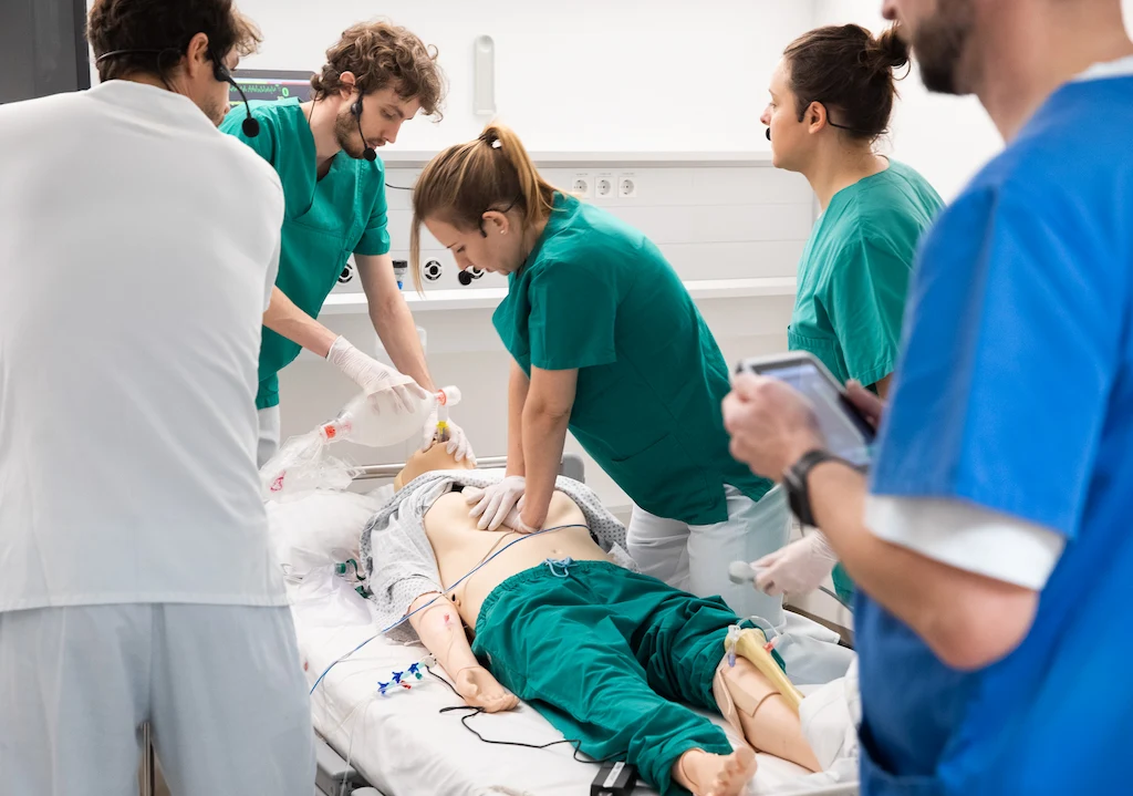 Medical trainees performing CPR on mannequin with chest compressions and ventilation, assisted by tablet monitoring.
