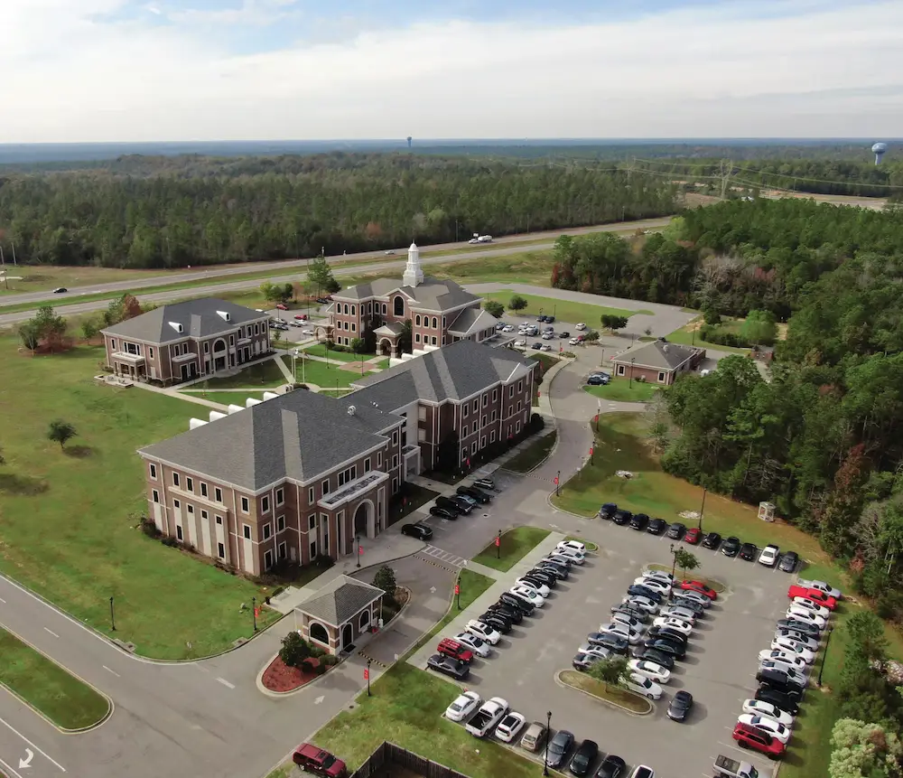 Aerial view of campus complex with brick buildings, cupola, parking lots, and surrounding forest landscape.
