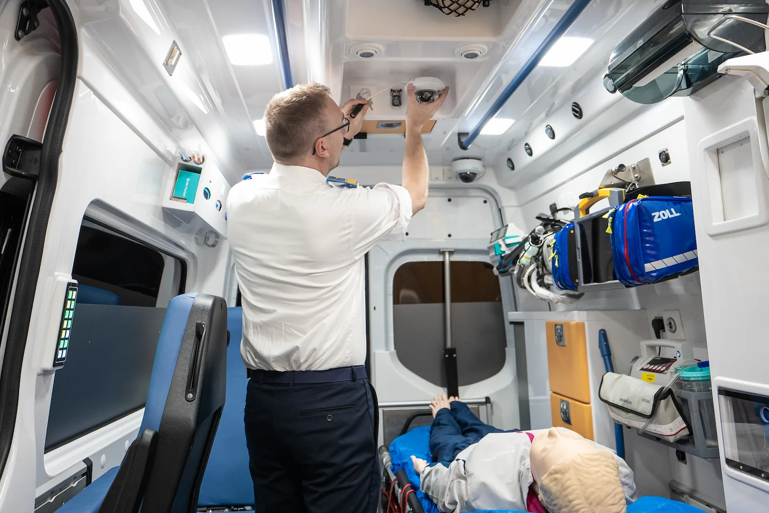 A paramedic adjusts controls on the ceiling inside an ambulance while a medical mannequin lies on a stretcher, surrounded by powerful SIMStation Mobile Systems for flexible simulation training and run video debriefings anytime.