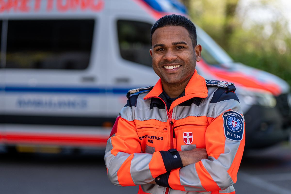 Paramedic in high-visibility uniform standing in front of an ambulance, smiling with arms crossed.